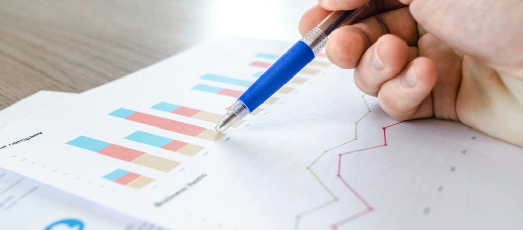 A close-up of a hand with a pen analyzing data on colorful bar and line charts on paper.